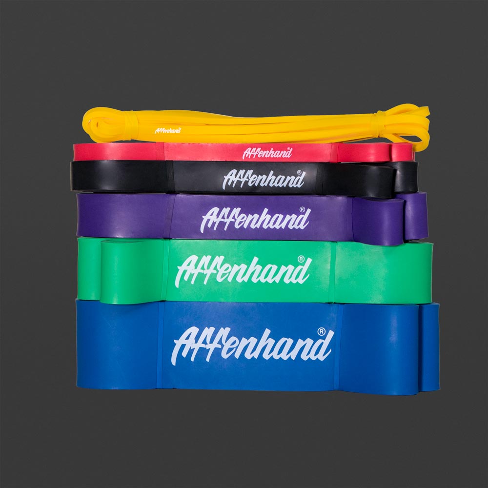Stapel farbiger Affenhand Widerstandsbänder – Gelb, Rot, Schwarz, Lila, Grün und Blau – mit weißem Logo auf dunklem Hintergrund.