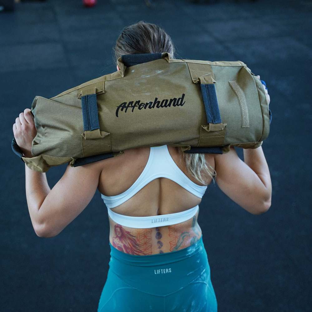 Athletin trägt einen braunen Affenhand Fitness Sandsack auf den Schultern, Rückenansicht mit weißem Sporttop und blaugrüner Leggings.