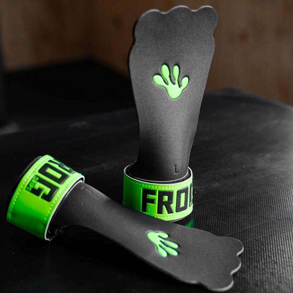 Zwei schwarze Frog Grips Base Grips 2.0 mit leuchtend grünen Logos und Handflächen-Design liegen auf einem dunklen Untergrund.
