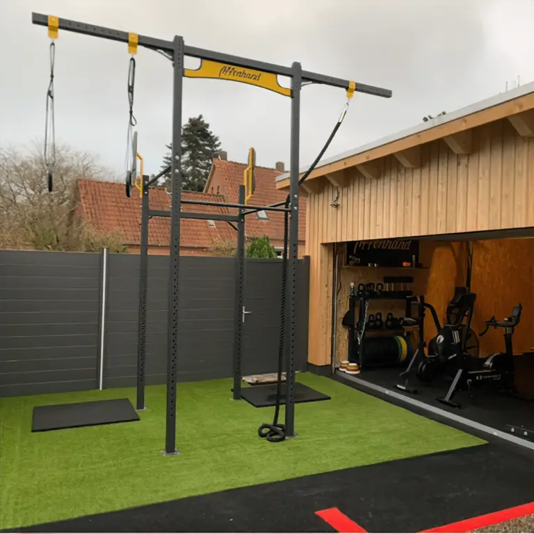 Affenhand-Rig mit Schlingentrainern auf einer Rasenfläche vor einem offenen Gym mit Ausdauergeräten.
