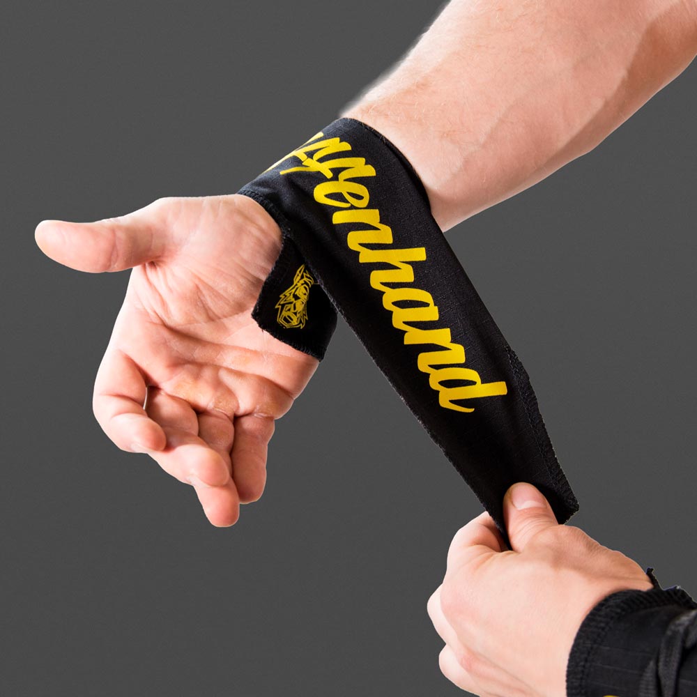 Eine Hand legt eine schwarze Affenhand Handgelenkbandage mit gelbem Schriftzug um das Handgelenk, auf grauem Hintergrund.