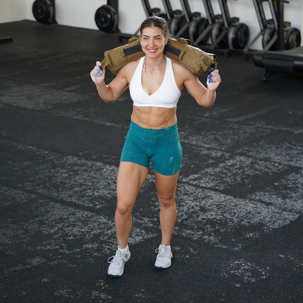 Athletin trägt den braunen Affenhand Fitness Sandsack auf den Schultern und lächelt während des Trainings in der Halle. Sie trägt ein weißes Sporttop und türkisfarbene Shorts.