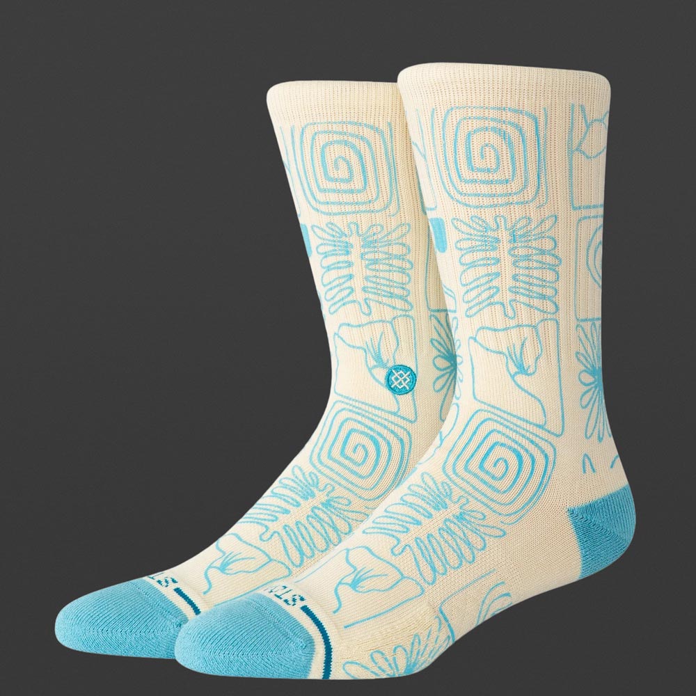 Ein Paar cremefarbene Stance Tested Everywhere Socken mit hellblauen Zehen, Fersen und geometrischem Muster aus Linien, Wellen und Blumen steht vor grauem Hintergrund.