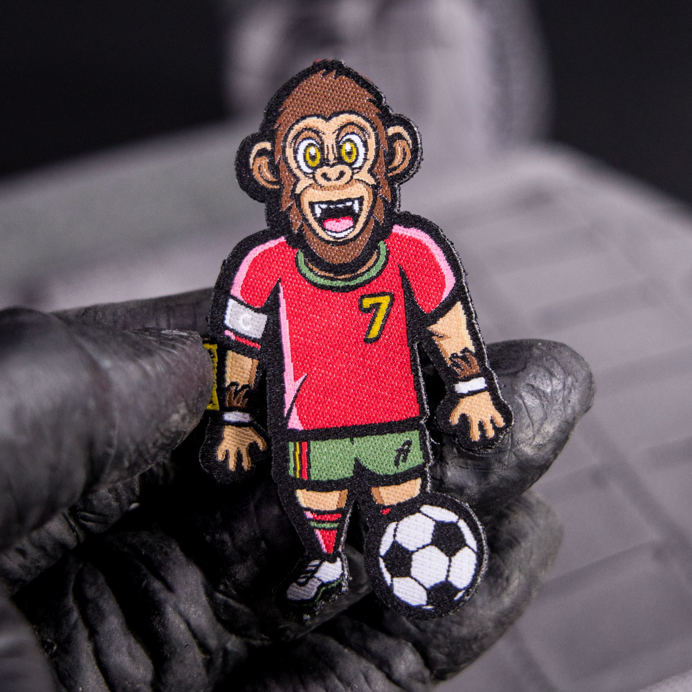 Hand hält einen Klett-Patch, auf dem ein dribbelnder Gorilla mit einem Fußball abgebildet ist.